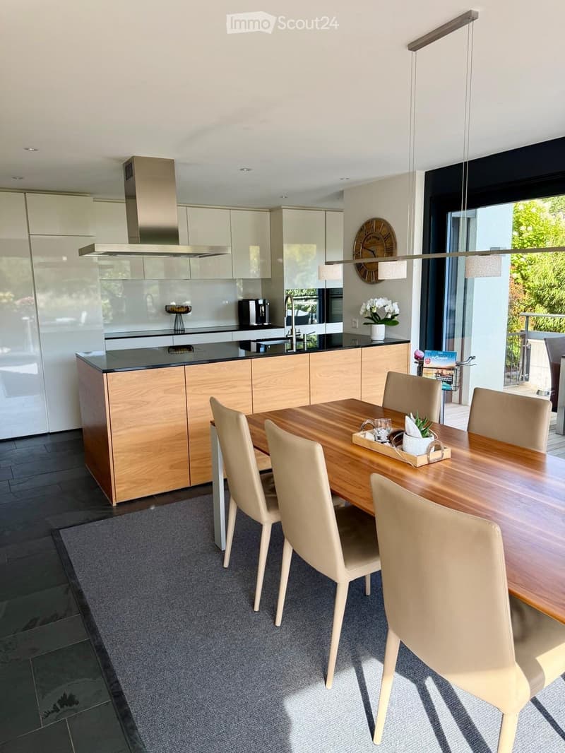 Modern kitchen with a wooden dining table, light-colored chairs, and a sleek kitchen island. Features include a stainless steel range hood, pendant lighting, and a vase with white flowers on the counter.