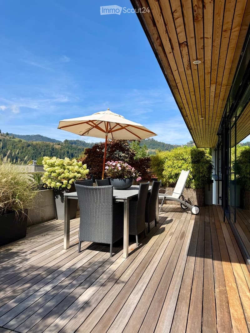 Wooden deck with outdoor furniture including a table, chairs, and an umbrella, surrounded by potted plants and a scenic view of hills and trees.
