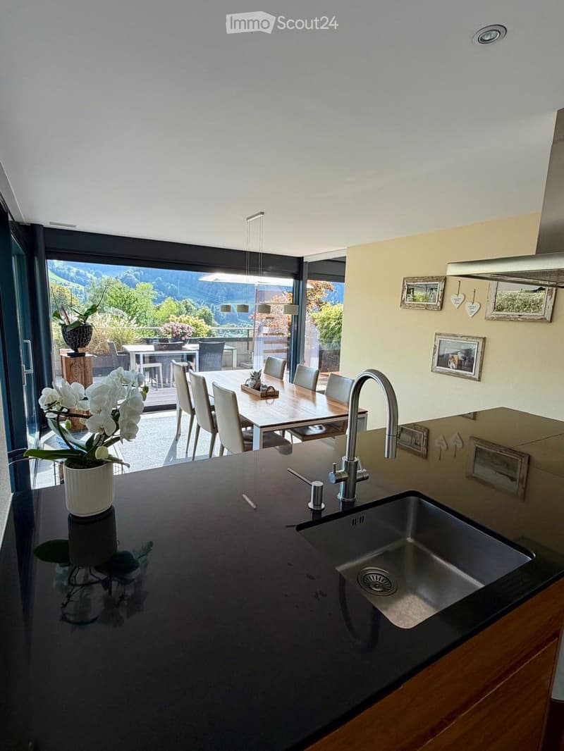 Modern kitchen with a dark countertop, stainless steel sink, and a dining area with a table and chairs. Large windows offer a scenic view of the landscape.