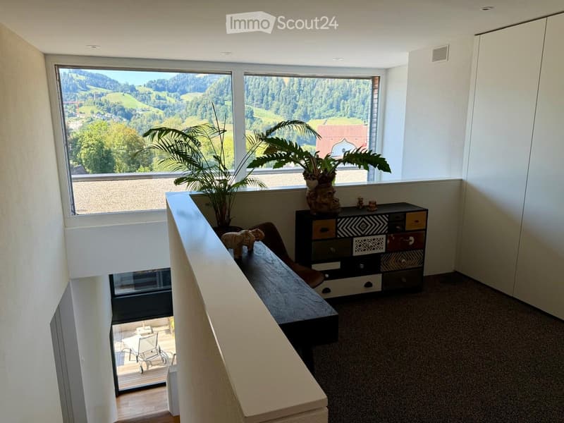 A modern, well-lit room with large windows offering a scenic view, featuring a sofa, a small table, a cabinet, and decorative items including a plant and a vase.