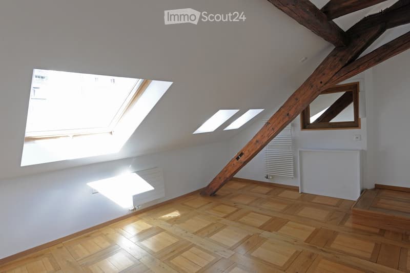 Bright attic room with wooden floor, exposed wooden beams, skylights, and a small mirror on the wall.