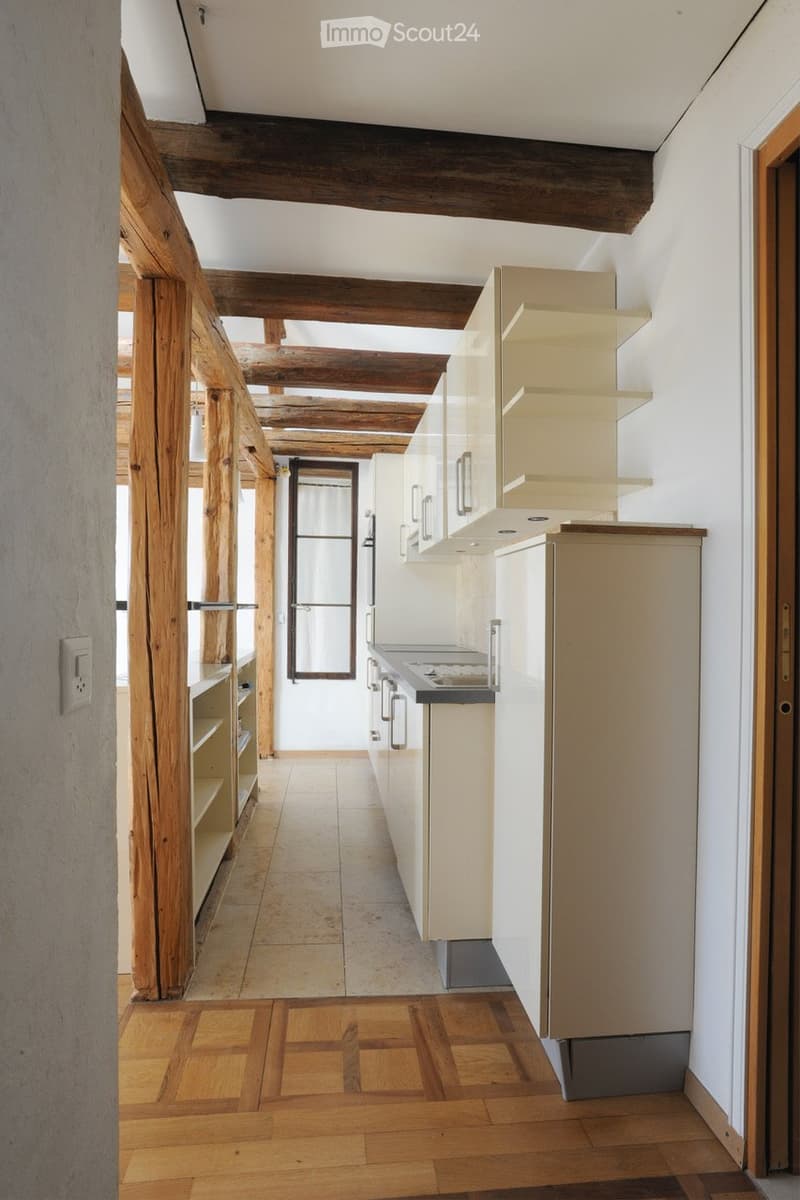 Modern kitchen with white cabinets, a stove, and a refrigerator. Exposed wooden beams on the ceiling and a mix of tiled and wooden flooring.
