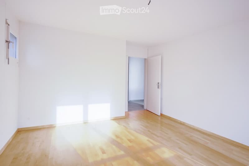 Empty room with white walls and wooden flooring, featuring a window and an open doorway leading to another space.