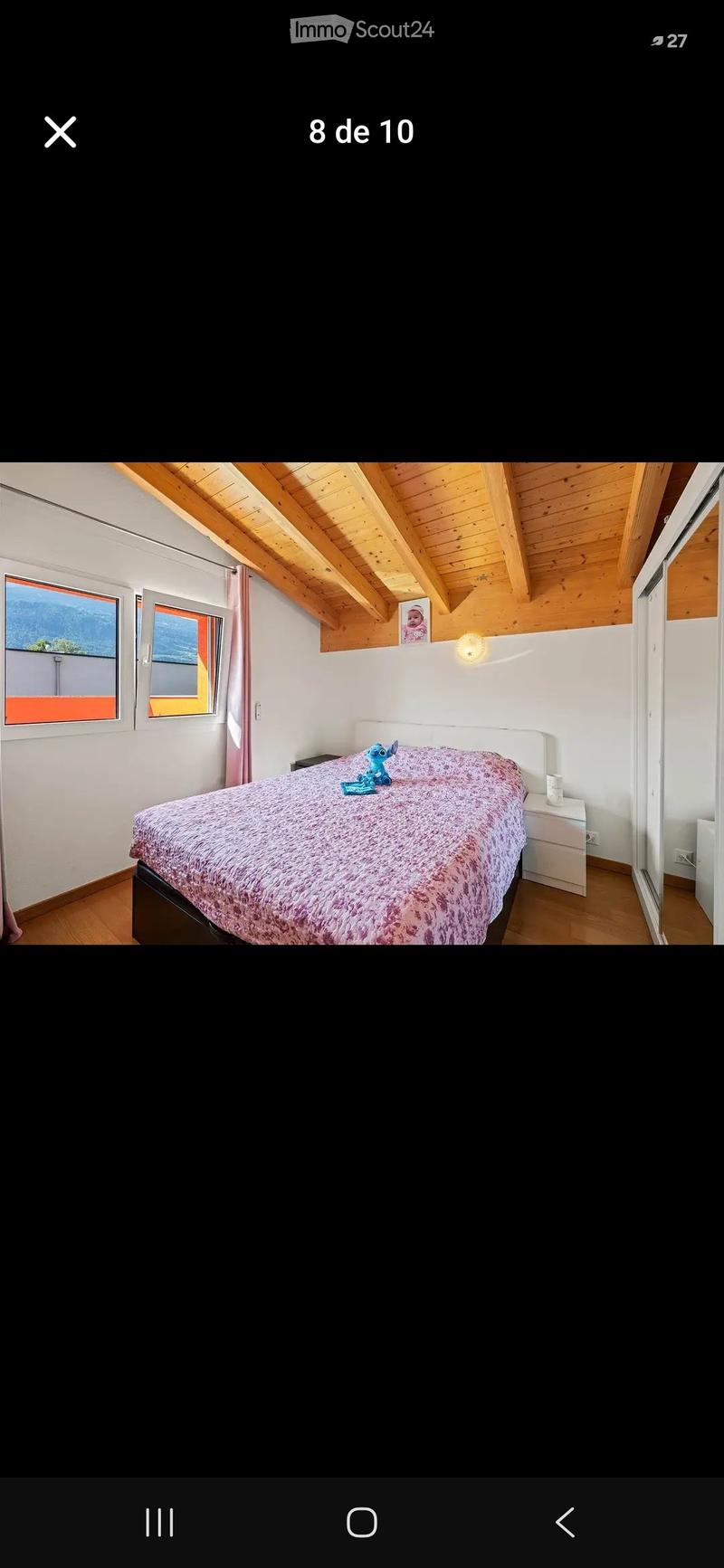 Cozy bedroom with wooden slanted ceiling, large windows with pink curtains, bed with pink patterned duvet, nightstand, and a small toy on the bed.