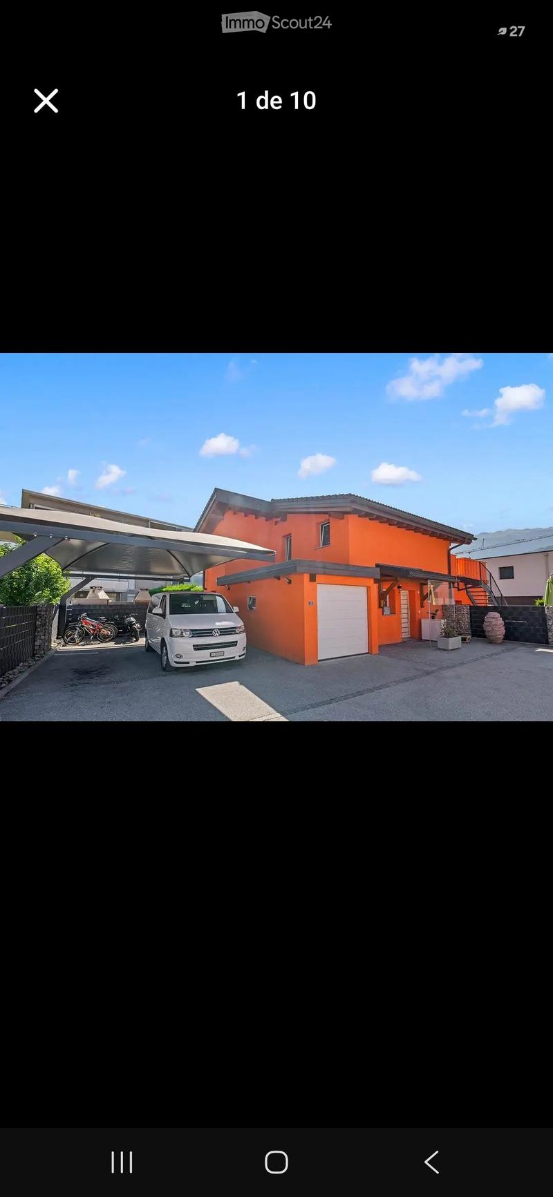 A modern two-story house with a bright orange facade, white garage door, and covered parking area. A white van is parked under the carport, and there are bicycles and motorcycles nearby.