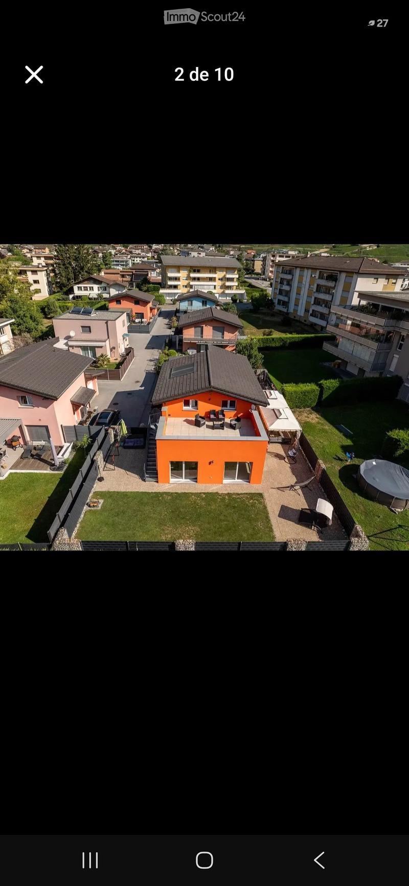 Aerial view of a residential area with multiple houses, including a prominent orange house with a terrace, surrounded by greenery and neighboring homes.