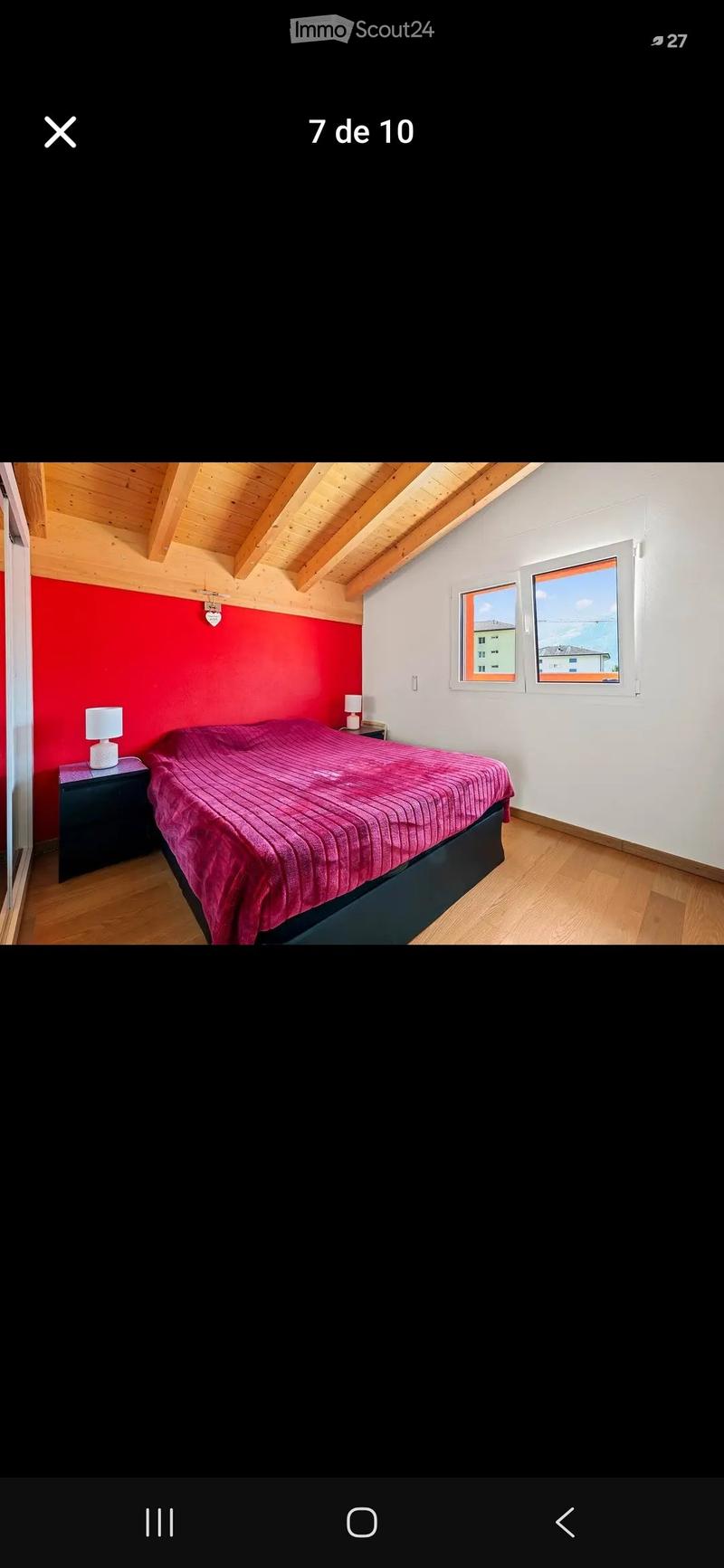Cozy bedroom with wooden slatted ceiling, red accent wall, and large windows offering a view. Features a neatly made bed with a pink duvet, two nightstands with lamps, and wooden flooring.