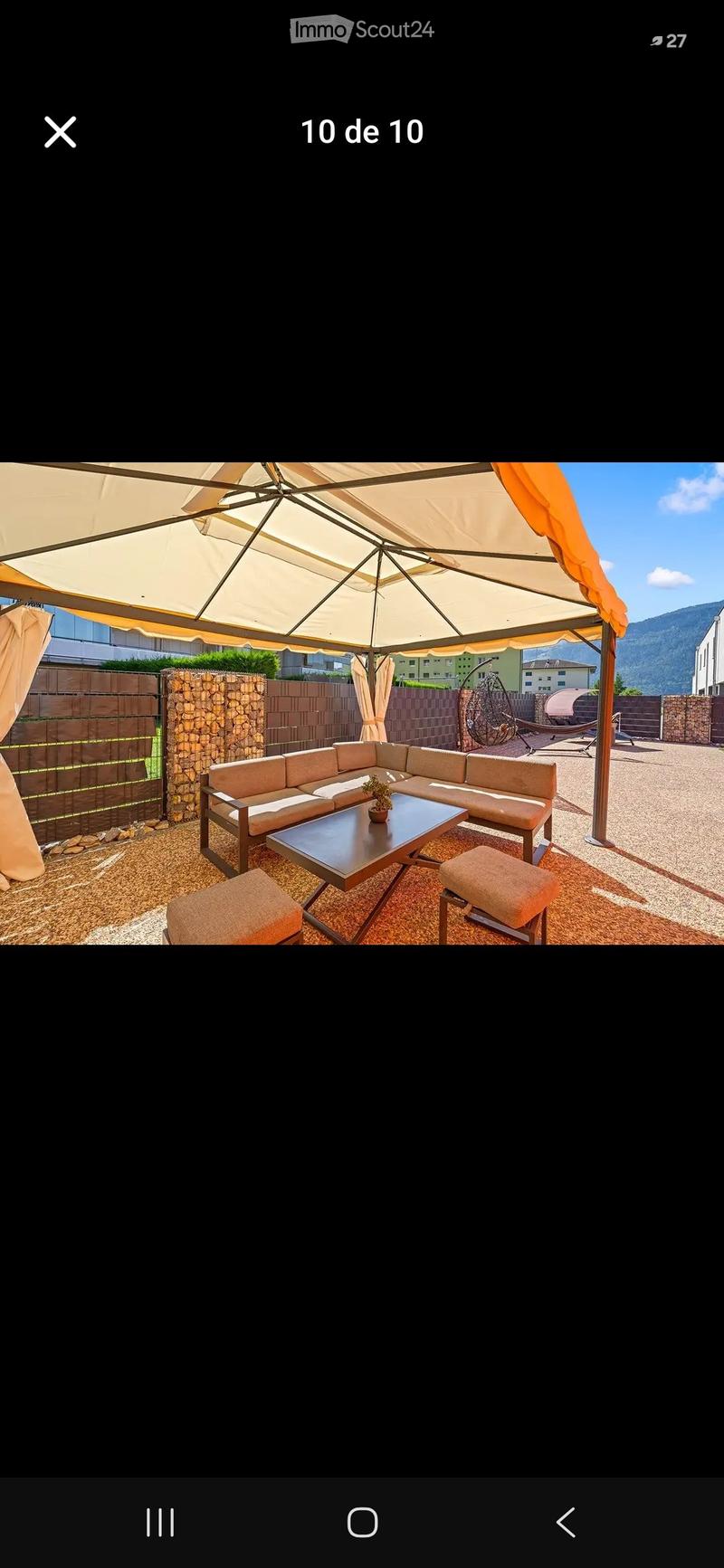 Outdoor patio with a beige sectional sofa, coffee table, two beige stools, and a large umbrella providing shade. The area is enclosed by a stone and wooden fence, with a view of mountains in the background.