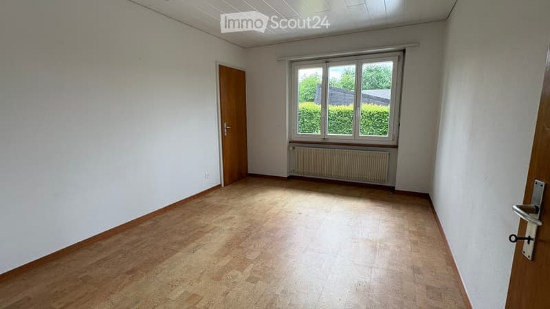 Empty room with wooden flooring, white walls, a window with blinds, and a radiator below it. There are two doors, one on each side of the room.