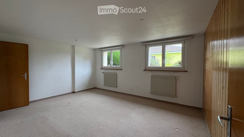 Empty room with white walls, carpeted floor, two windows, and radiators. No furniture or equipment present.