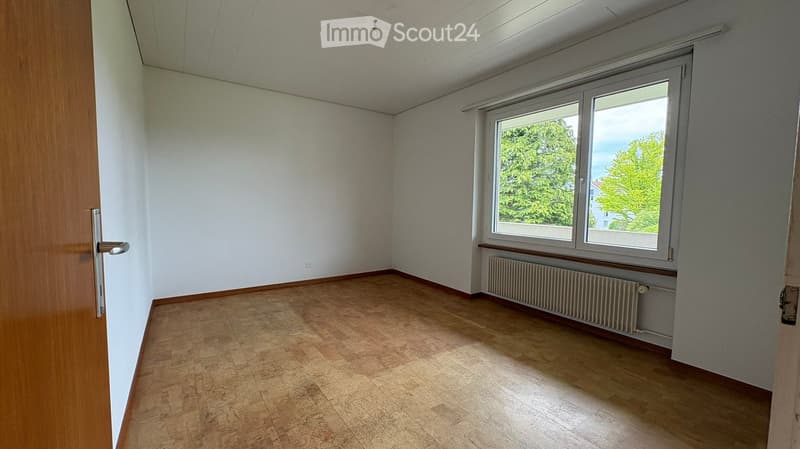 Empty room with wooden flooring, white walls, and a large window with a view of greenery outside. A radiator is placed under the window, and a door is visible on the left side.