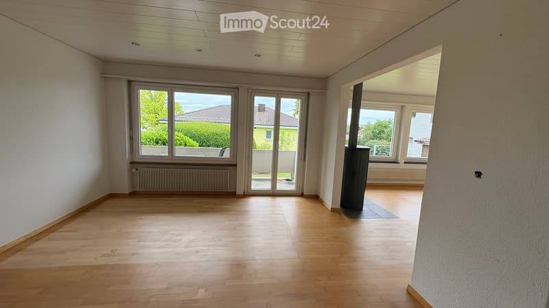 Empty room with wooden flooring, white walls, large windows, and a central heating column.