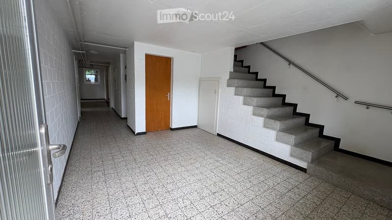 Empty hallway with tiled floor, white walls, a staircase with gray steps, and a wooden door.