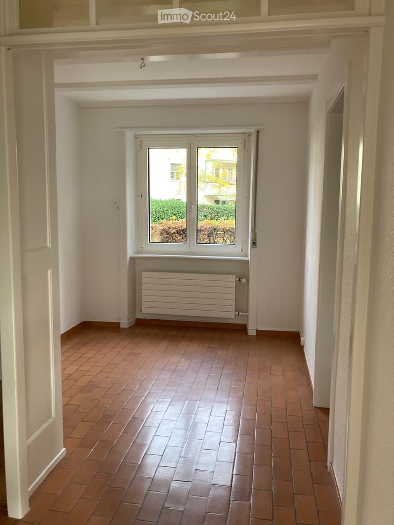Empty hallway with tiled floor, white walls, and a window at the end showing greenery outside.