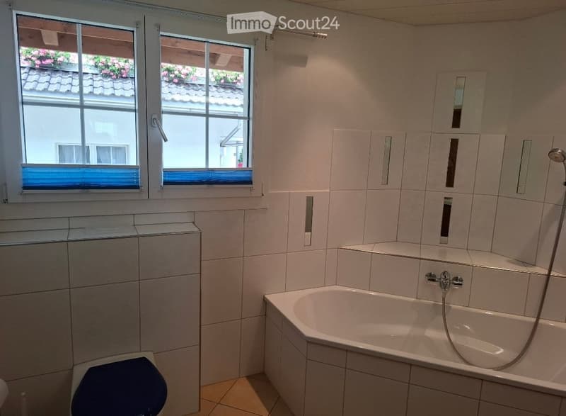 Modern bathroom with white tiled walls, a bathtub, toilet, and a large window offering a view of a tiled roof and flowers outside.