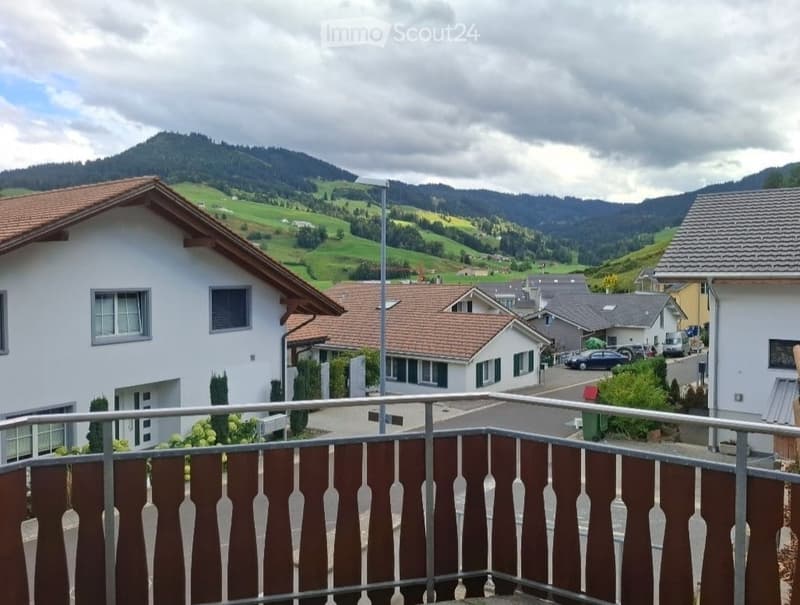 Balcony overlooking a scenic countryside with houses, green fields, and distant mountains.