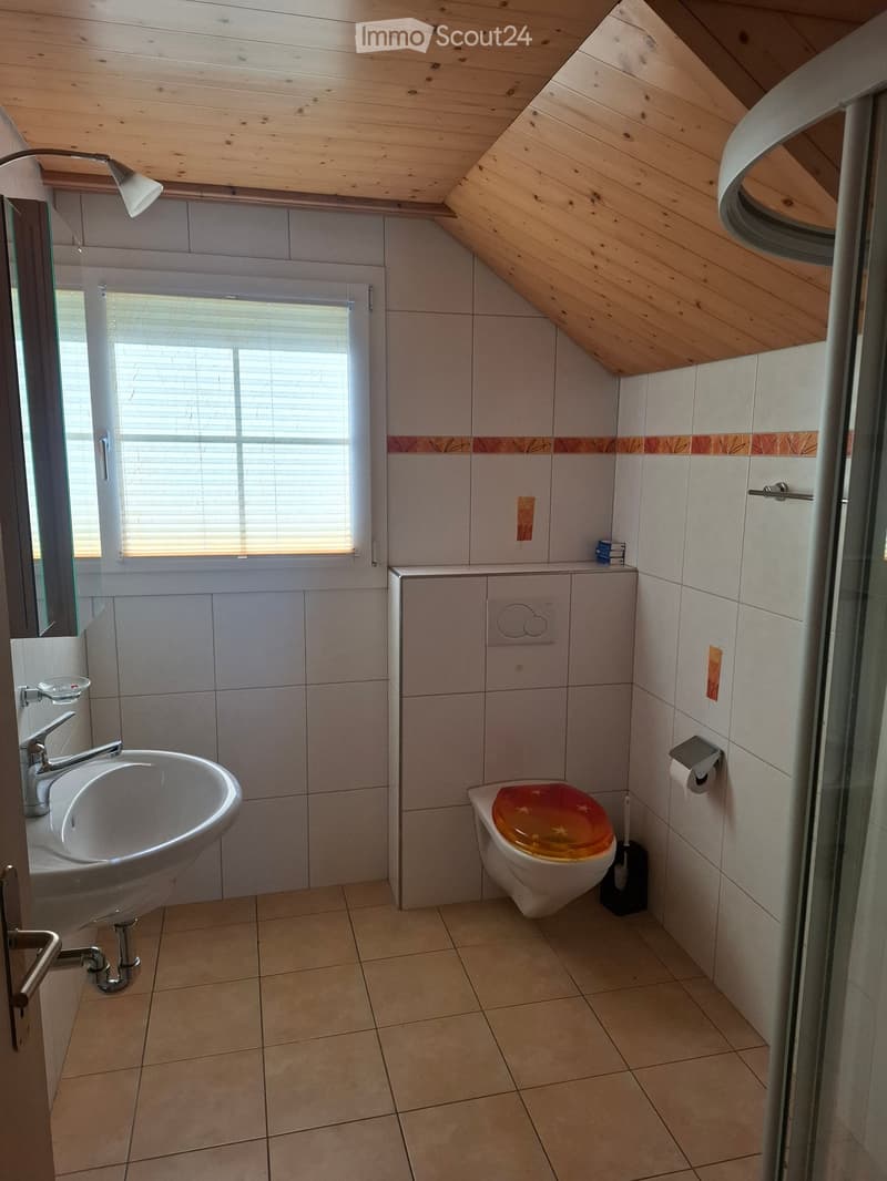 Bathroom with a sink, toilet, and a window with blinds. The room features tiled walls and a wooden slanted ceiling.