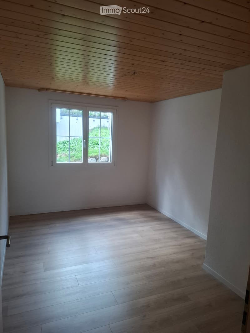 Empty room with wooden flooring, white walls, and a large window letting in natural light.