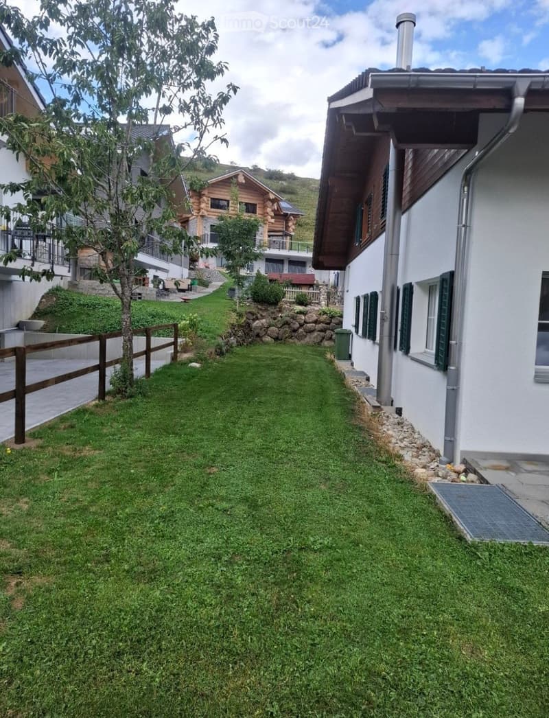 A well-maintained lawn area bordered by a wooden fence on one side and a stone retaining wall on the other, with a small tree and a few bushes. A white house with green shutters is visible on the right, and a log cabin-style house is seen in the backgroun