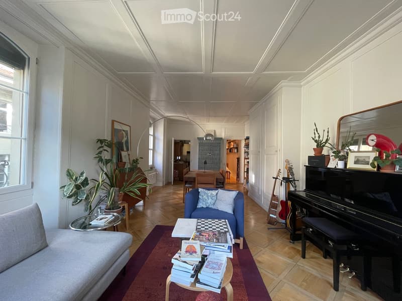Living room with a grey sofa, coffee table, small round stool, books, piano, guitar, dining area with table and chairs, and plants.