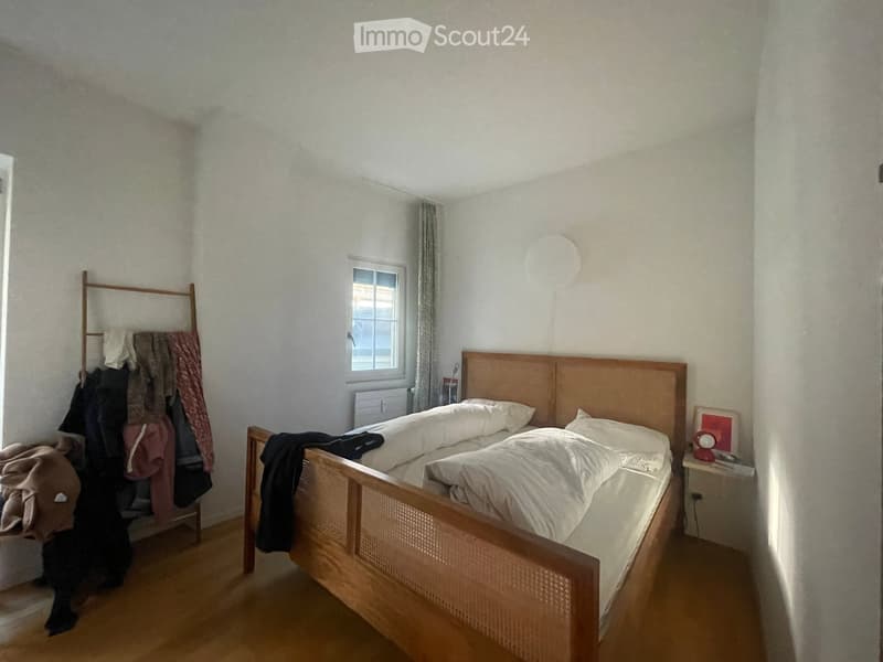 A small, well-lit bedroom with a wooden bed, white bedding, a nightstand with a lamp, a window with curtains, and a clothing rack with folded clothes.