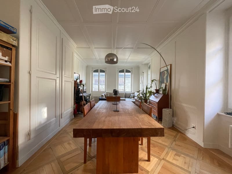 Bright, open-plan living and dining area with wooden flooring, large windows, a long wooden dining table, chairs, a sofa, a guitar, a piano, a bookshelf, a floor lamp, and a plant.
