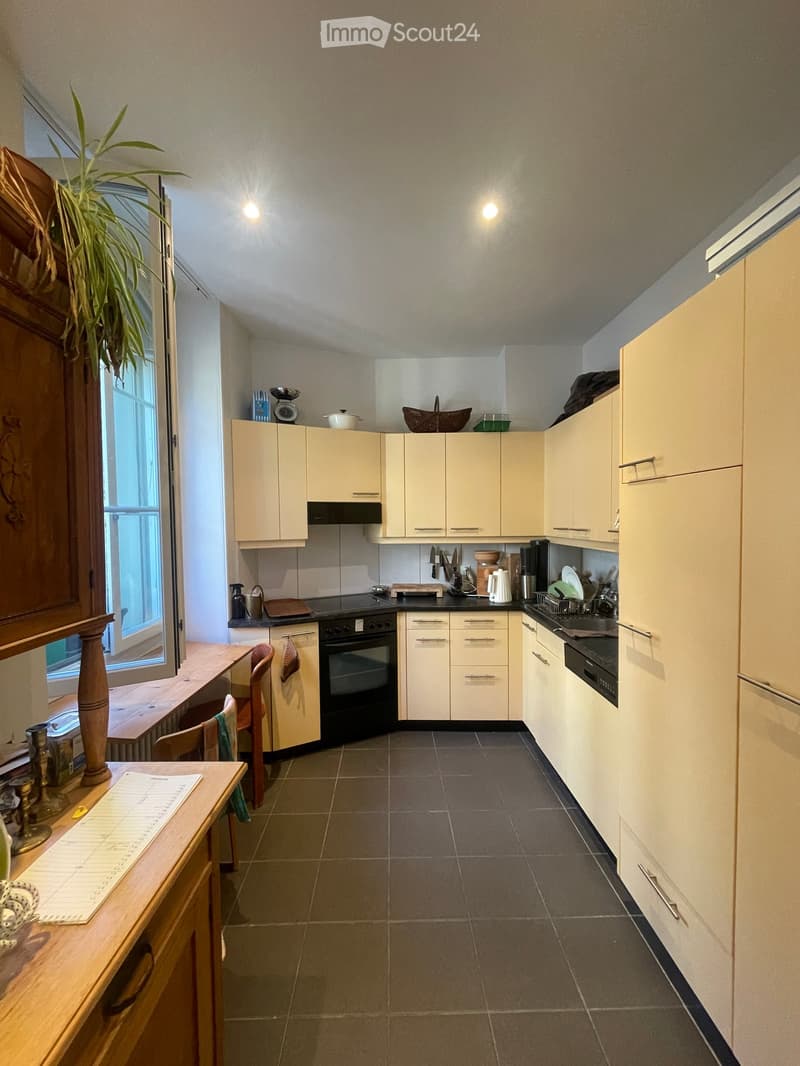Modern kitchen with light-colored cabinets, dark tiled floor, and various appliances including an oven, sink, and dish rack. A small wooden table with chairs is positioned near a window, and a potted plant adds a touch of greenery.