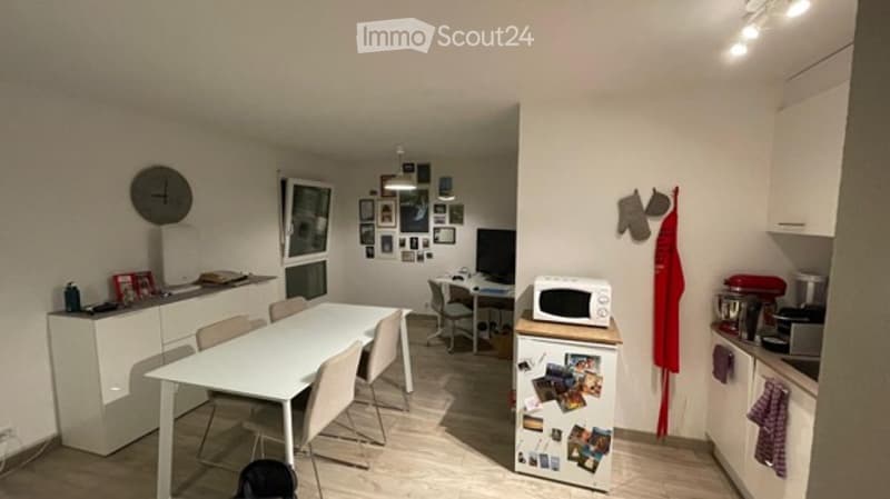 A modern, open-plan living and dining area with a white dining table and chairs, a small kitchen area with cabinets, a refrigerator, microwave, and various kitchen appliances, a desk with a laptop, and a wall decorated with picture frames and a clock.