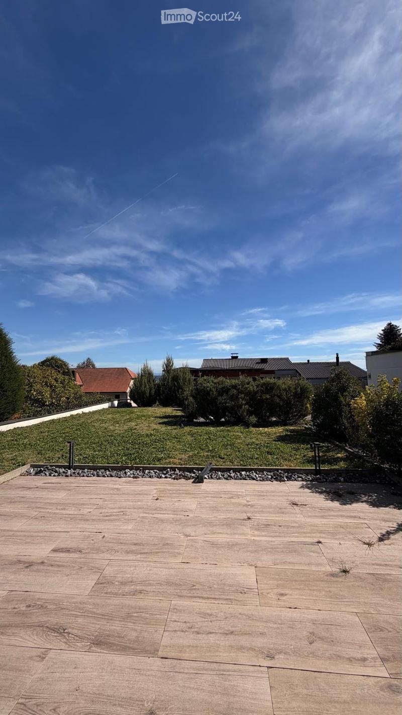Wooden deck with a gravel border, surrounded by greenery and distant houses under a clear blue sky.