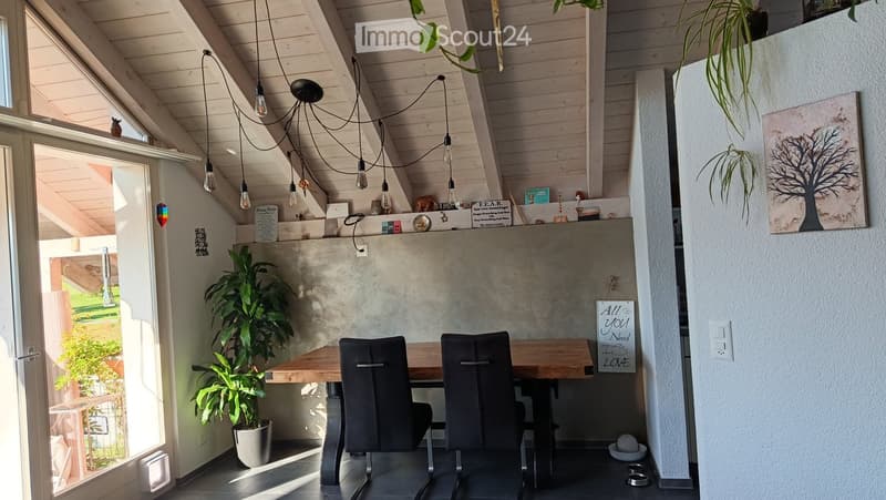 Bright, modern dining area with wooden table, two chairs, potted plants, and a painting on the wall. Exposed wooden beams on the ceiling and pendant lights.