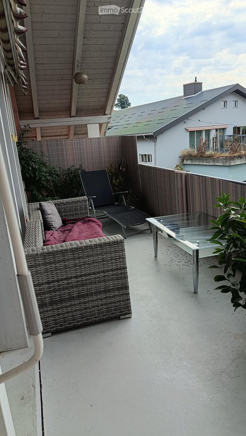Balcony with a wicker sofa, a black lounge chair, a glass table, and a small plant. The balcony is enclosed with a wooden fence and has a view of neighboring buildings.