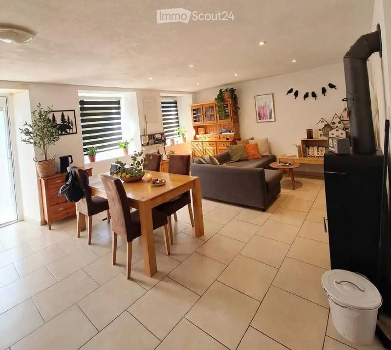 Bright living and dining area with tiled flooring, large windows with blinds, a grey sofa, a wooden dining table with chairs, a wooden cabinet, and decorative plants.