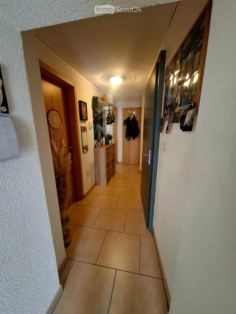 A narrow hallway with tiled flooring, beige walls, and several doors leading to different rooms. There are small shelves and decorations visible on the walls.