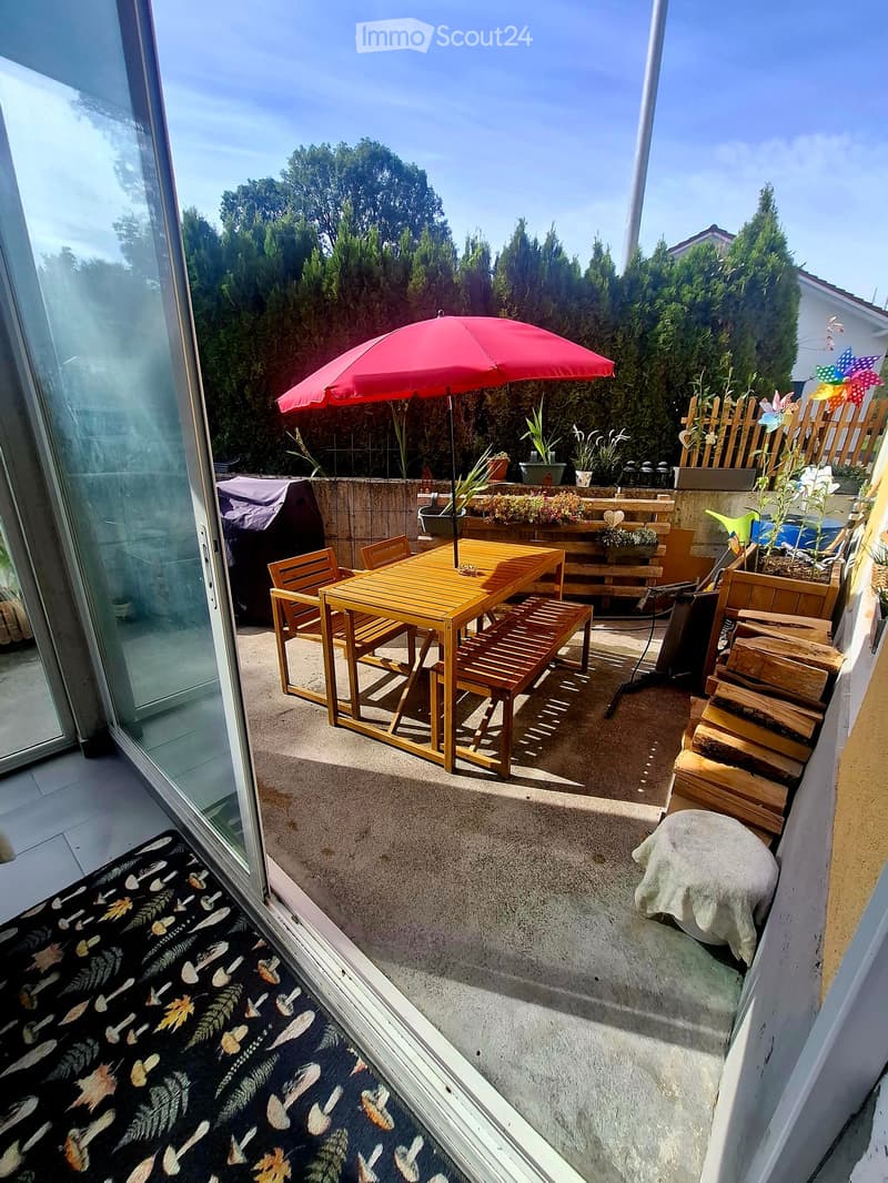 Balcony with a wooden table, chairs, and a red umbrella, surrounded by potted plants and a wooden fence.