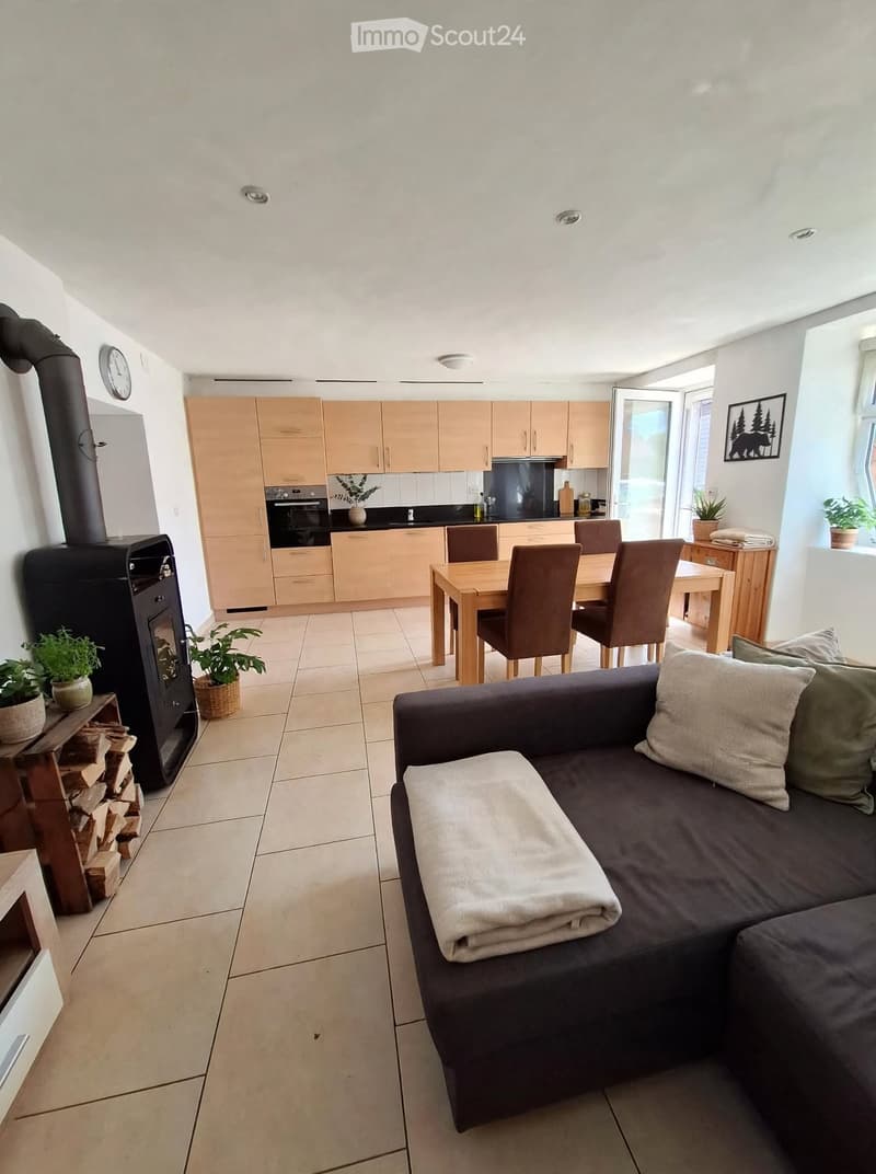 Open-plan living and dining area with a dark grey sofa, a wooden dining table with chairs, a kitchen area with light wooden cabinets, and a black wood-burning stove.