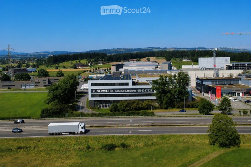 Aerial view of a modern industrial complex with multiple buildings, parking lots, and a highway running nearby. The main building features a sign advertising office space for rent.