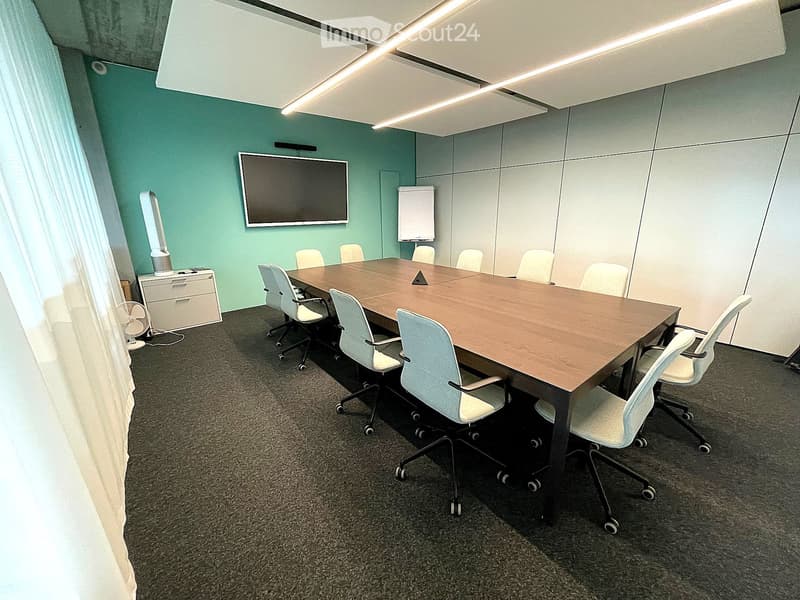 Conference room with a long wooden table surrounded by white chairs, a mounted screen, a fan, and a small cabinet with drawers.