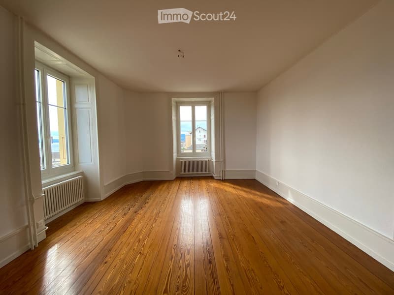 Empty room with wooden flooring, white walls, and large windows offering a view of the outside. Radiators are visible beneath the windows.