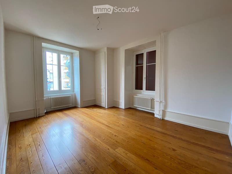 Empty room with wooden flooring, white walls, two windows, and radiators beneath them.