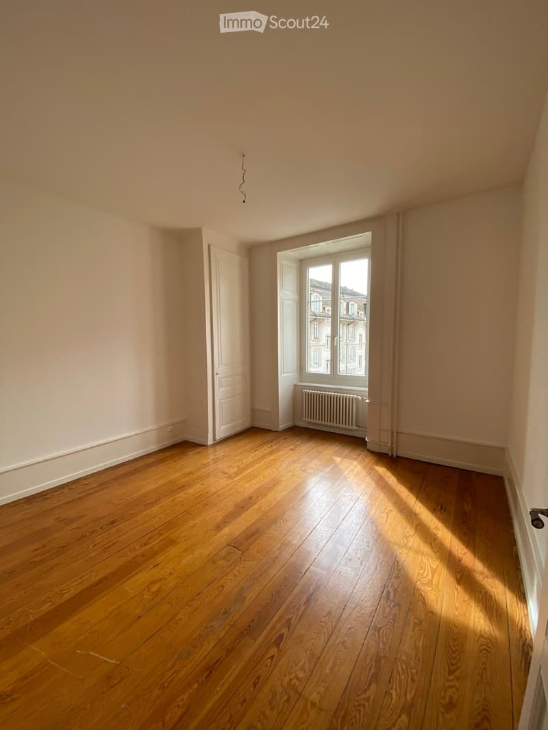 Empty room with wooden flooring, white walls, a single door, and a window with a radiator below it. Natural light streams in from the window, illuminating the space.