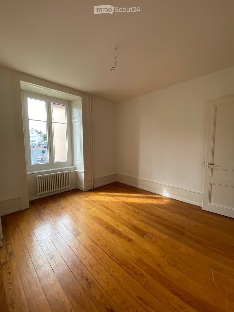 Empty room with wooden flooring, white walls, and a large window with a radiator below it. The room has a door on the right side and is well-lit with natural light.