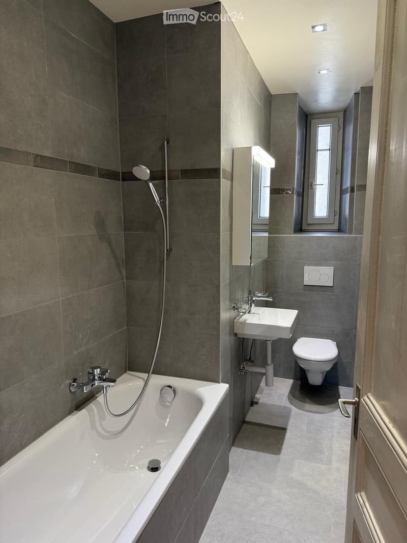 Modern bathroom with a bathtub, showerhead, sink, and toilet. Grey tiled walls and floor, sleek fixtures.