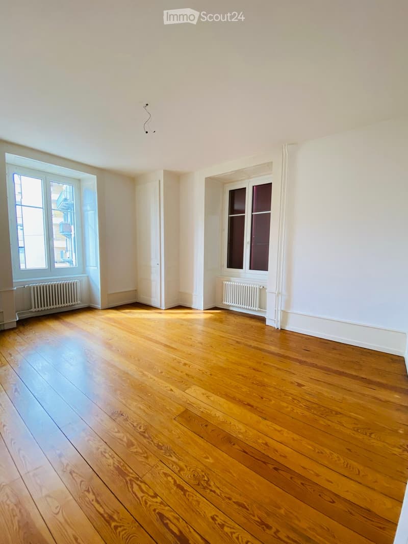 Empty room with wooden flooring, white walls, and two windows with radiators beneath them. The room has a door leading to another space.