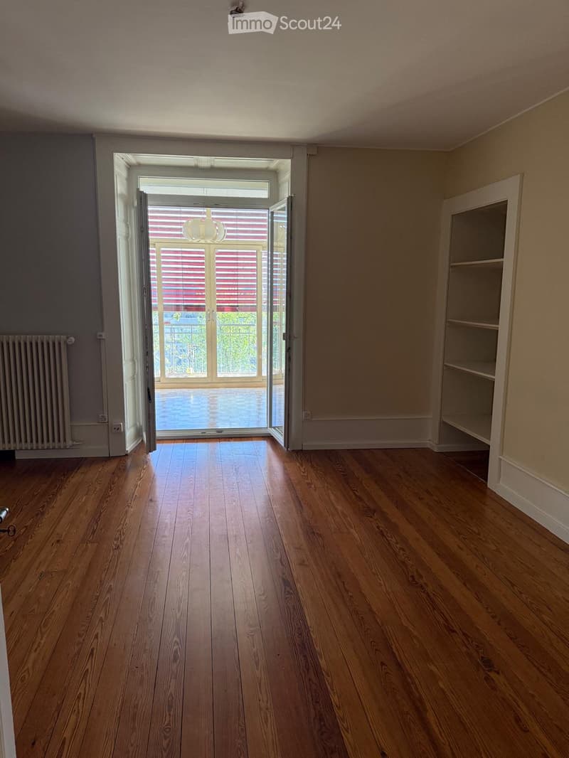 Empty room with wooden flooring, white walls, built-in shelving unit, and a large open doorway leading to a balcony or outdoor area.