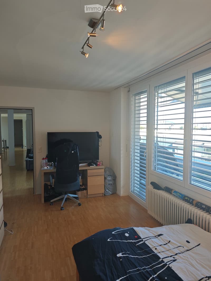 Bright bedroom with wooden flooring, large windows with blinds, a bed with a blue and white patterned duvet, a desk with a chair, and a TV screen on a stand.