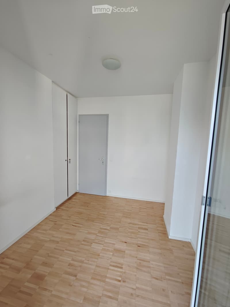 Empty room with light wooden flooring, white walls, and a single door. The ceiling has a round light fixture.