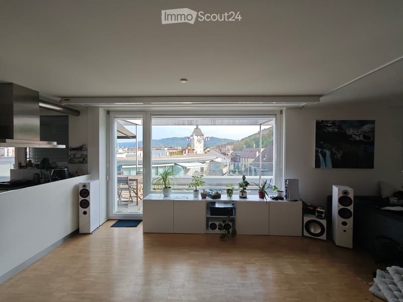 Bright living room with wooden flooring, large windows offering a scenic view, a sofa, coffee table, potted plants, and a kitchen area with appliances.