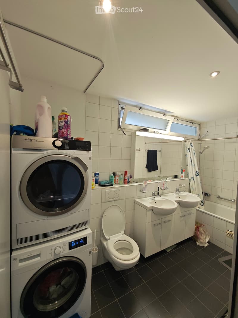Modern bathroom with a stacked washer and dryer, toilet, double sink, bathtub, and mirror. Cleaning supplies are stored on top of the washer.