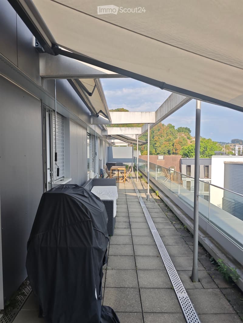 Balcony with covered area, outdoor furniture, and glass railing, overlooking a landscaped area with trees and neighboring buildings.