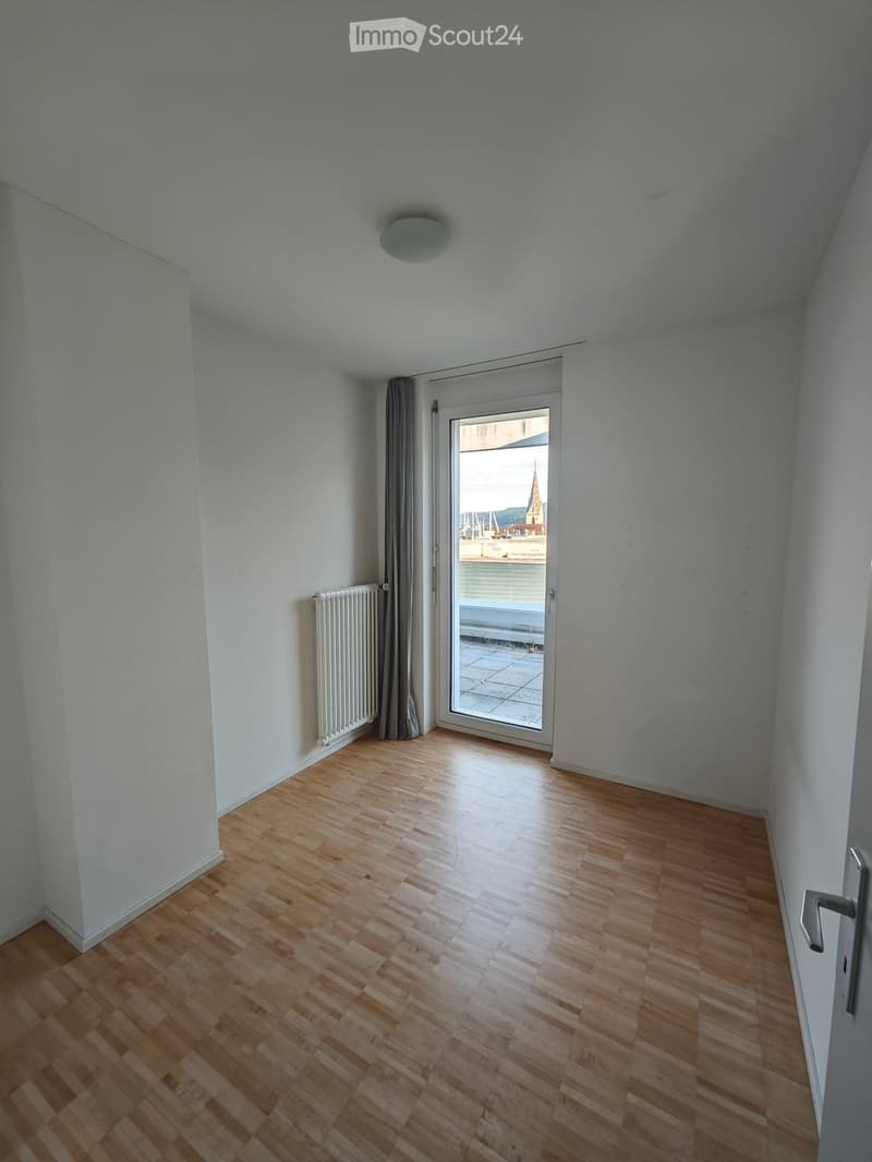Empty room with wooden flooring, white walls, a radiator, and a sliding glass door leading to a balcony with a view of a cityscape.